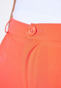 Heldere oranje broek met een gladde stoftextuur, een knoop aan de voorkant met vier gaatjes en zijzakken met subtiele stiksels.
