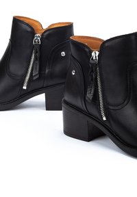 Botas de cuero negras hasta el tobillo con un bajo tacón cuadrado, cremalleras laterales y una textura suave. Interior forrado con cuero marrón. Costura decorativa visible.