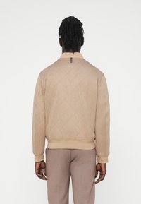 Μπεζ καπιτονέ μπουφάν τύπου bomber με ριμπ γιακά και μανσέτες, που διαθέτει διακοσμητικό φερμουάρ στο πίσω μέρος. Φοριέται με μπεζ παντελόνια.