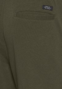 Olivgrüner Stoff mit einer weichen Textur; verfügt über eine Gesäßtasche und ein kleines schwarzes Etikett mit weißem Text.