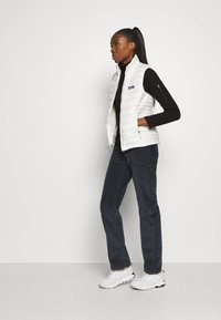 Witte geïsoleerde vest met een hoge kraag, ritszakken en een logo-opdruk, gecombineerd met donkere broek en witte sneakers. Soepele textuur.