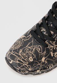 Baskets noires avec des motifs graphiques beiges complexes. Tige en texture lisse, lacets noirs et détails perforés près de la zone des orteils.
