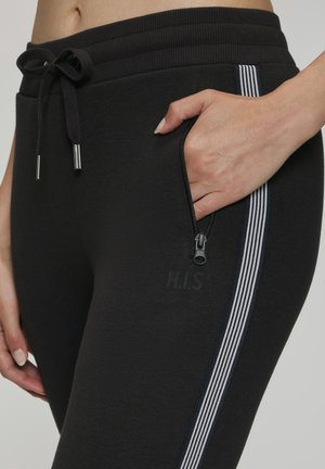 Hand with manicured nails in side zippered pocket of black joggers with white striped side detail and drawstring waistband.
