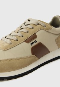 Sapatilha bege feita de têxtil e camurça, apresentando um detalhe em pele castanha e o logótipo "BOSS". Cadarços planos e sola de borracha preta texturizada.