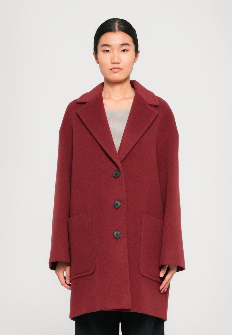 Manteau en laine oversize bordeaux avec un col cranté, cinq boutons noirs et deux poches latérales ; texture lisse et coupe décontractée.