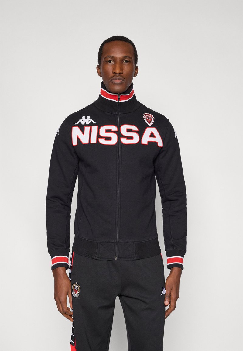 Svart jacka med dragkedja och hög krage, röda och vita accenter, stora bokstäver "NISSA" och Kappa-logotyp på ärmen. Har ribbstickade muddar.