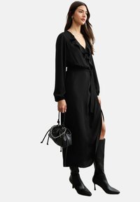 Robe noire à manches longues avec un col à volants, une ceinture à la taille et une fente sur le côté. Associée à des bottes à talons noires et un petit sac noir.