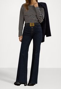 Camisa de rayas de manga larga combinada con pantalones acampanados de mezclilla oscuros de cintura alta, que cuentan con un cinturón ancho con un emblema dorado y tres botones en la parte frontal.