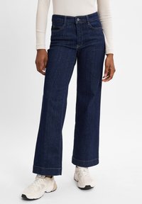 Høy midje, mørkeblå denimjeans med vid ben, med fem lommer og subtile sømdetaljer, kombinert med lyse sportsko.