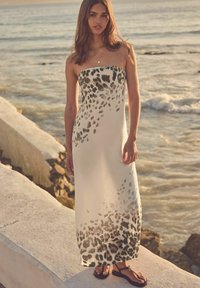 Abito lungo senza spalline in tessuto bianco trasparente con una stampa leopardata nera, caratterizzato da una silhouette fluida e sandali con suola piatta.