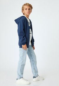 Sudadera con cremallera de color azul marino, camiseta gráfica blanca, jeans de color azul claro y zapatillas blancas con detalles grises. Tejido suave y corte relajado.