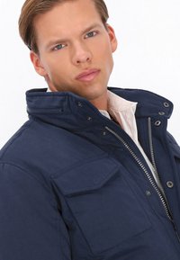 Mørkeblå zip-up jakke med høj krave, to brystlommer og fremtrædende syninger. Har knaplukninger og en robust tekstur.
