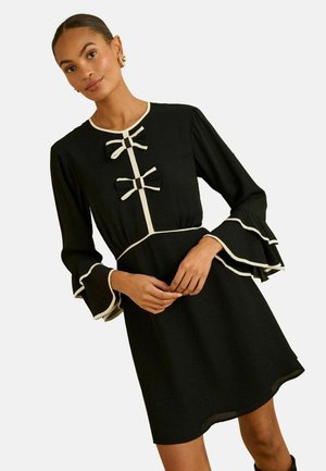 Melna kleita ar garām volānu piedurknēm, baltu kontrastējošu apdari un divām lokām priekšpusē. Gluda auduma, ceļgalu garuma dizains.