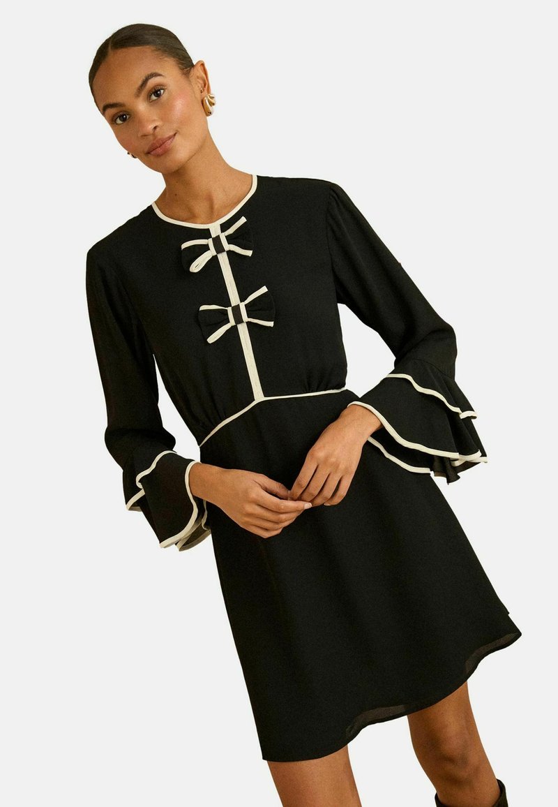 Černé šaty s dlouhými, volánkovými rukávy, bílým kontrastním lemováním a dvěma mašlemi vpředu. Hladká látka, délka ke kolenům.
