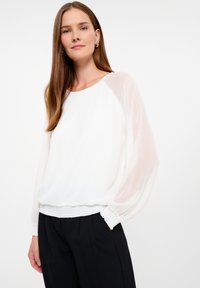 Witte blouse met doorschijnende, puffy mouwen en een ronde halslijn; textuurstof met een verzamelde zoom; gedragen met zwarte broek.
