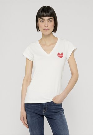Frau mit kurzen dunklen Haaren, die ein weißes V-Ausschnitt-T-Shirt mit einem roten Flammenherz-Logo und blaue Jeans trägt, steht mit einer Hand in der Tasche.