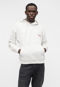 Ανοιχτό γκρι φούτερ με κουκούλα από μαλακό ύφασμα, διαθέτει εμπρόσθια τσέπη και ρυθμιζόμενη κουκούλα με κορδόνι. Έχει κόκκινο κεντημένο λογότυπο στο στήθος. Φορεμένο με σκούρο τζιν.