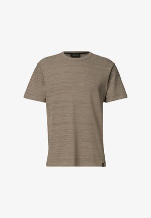 Homme aux cheveux courts et foncés portant un t-shirt marron clair à manches courtes et col rond, debout devant un fond clair uni.