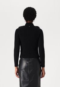 Person med kort, krøllet hår iført en sort ribbet sweater og sort lædernederdel, vist bagfra mod en enkel baggrund.