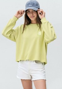 Felpa oversized verde chiaro con una piccola grafica sul lato sinistro del petto, abbinata a pantaloni shorts in denim bianco strappati e un cappellino da baseball grigio.