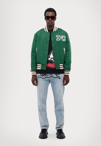 Grønn bomberjakke med stripete mansjetter, grafisk svart t-skjorte, lyseblå jeans og svarte sko. Modellen har på seg solbriller.