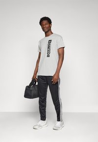 Grå t-skjorte med svart "BOXING" trykk på venstre side, kombinert med svarte treningsbukser med hvite striper. Modell holder en svart treningsbag.