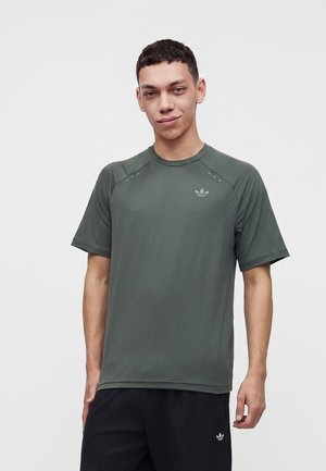 Mladý muž v tmavozelenom tričku Adidas s krátkym rukávom a čiernych nohaviciach Adidas stojí pred jednoduchým svetlým pozadím.