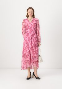Robe maxi fleurie rose avec manches longues, design enveloppant et jupe à étages. Portée avec des talons noirs et tenant un sac à main blanc.