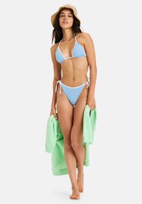 Lichtblauwe bikini met witte zoom, met een driehoektop en een strikzijde broekje. Model houdt een muntgroene sweatshirt vast.