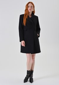Cappotto doppiopetto nero con grandi bottoni, maniche lunghe e tasche laterali, caratterizzato da una silhouette strutturata e un tessuto testurizzato.