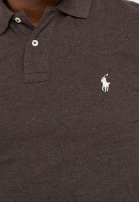 Brun polotröja med en texturerad stickning, prydd med en vit broderad logotyp på bröstet och en treknappsslutning.