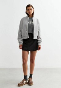 Szara polarowa bomberka z ściągaczami, założona na szary sweter z napisem „HER” w jasnym kolorze. Czarna spódnica w paski i trampki w leopardzi wzór.