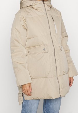 Beige gewatteerde jas met rits aan de voorkant, borstzak met rits, twee klepzakken met knopen en een capuchon, gedragen met een blauwe spijkerbroek.