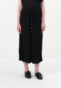Black wide-leg trousers made of flowy fabric with a drawstring waist, featuring gold accents and a soft texture. Paired with black shoes.