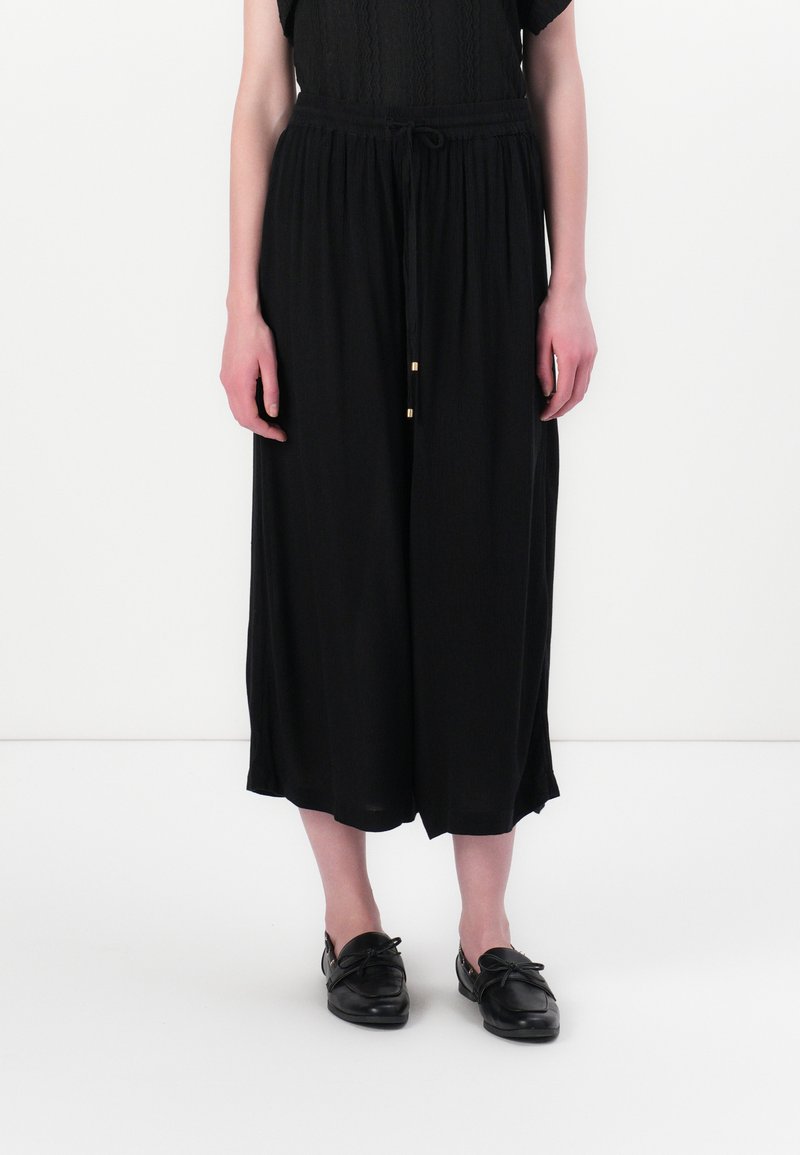 Black wide-leg trousers made of flowy fabric with a drawstring waist, featuring gold accents and a soft texture. Paired with black shoes.