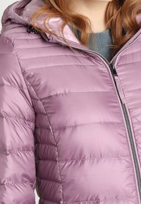 Lys lilla pufferjakke med vandret quiltning, en lynlåslukning foran og en aftagelig hætte. Den har en glat, blank tekstur og kontrasterende lynlås.