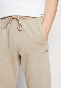 Beiga mjukisbyxor gjorda av mjuk bomull, med dragsko i midjan, sidofickor och en diskret broderad logga i mörkrosa.