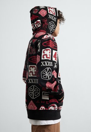 Schwarzer Kapuzenpullover mit roten und weißen grafischen Mustern, die Logos und Zahlen zeigen. Das Material scheint weich zu sein; er hat eine Kordelzugkapuze und einen gerippten Saum.
