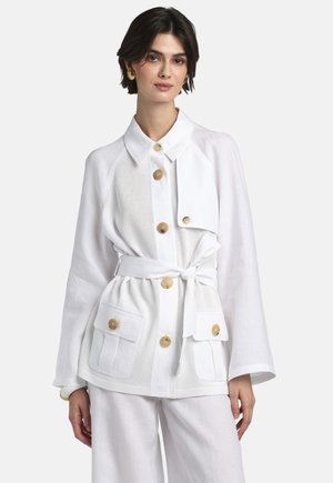 Donna che indossa una giacca bianca con cintura, grandi bottoni beige e tasche frontali, abbinata a pantaloni bianchi a gamba larga e orecchini dorati.