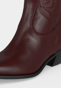 Καφέ δερμάτινη καουμπόικη μπότα με μυτερή μύτη, διακοσμητικές ραφές και χαμηλό κεκλιμένο μαύρο τακούνι σε λευκό φόντο.