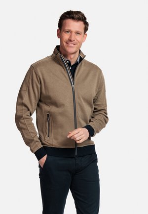 Bruine zip-up jas met een textuurafwerking, zwarte manchetten en tailleband, twee zijzakken met ritsen. Gedragen over een zwart overhemd met kraag.