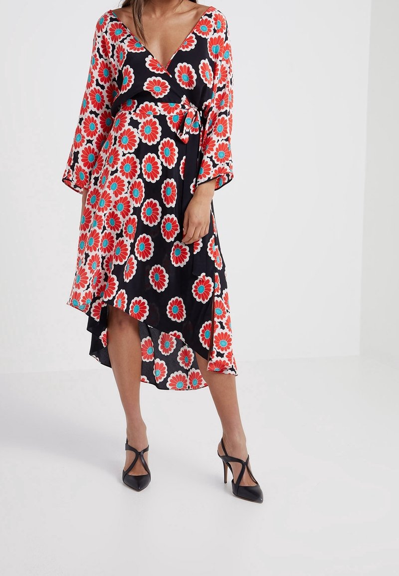Femme portant une robe portefeuille noire à grosses fleurs rouges et bleues, manches trois-quarts et ourlet asymétrique, associée à des talons noirs.