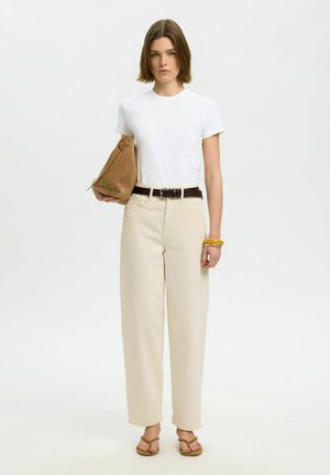 Kvinne med kort brunt hår, iført hvit t-skjorte, beige høytlivsbukser, brunt belte, sandaler, gullarmbånd, og holder en lysebrun semsket veske ved siden.