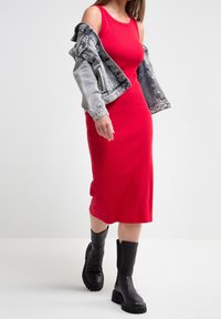 Czerwona, prążkowana sukienka midi o dopasowanym kroju, w połączeniu z jasnoniebieską kurtką dżinsową. Model nosi czarne botki za kostkę na grubej podeszwie.