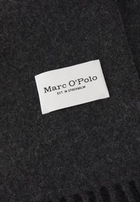 Sötét szürke gyapjú s�ál puha tapintással és rojtolt szélekkel, amelyen egy fehér címke található, ami a következő szöveget tartalmazza: "Marc O’Polo EST. IN STOCKHOLM."