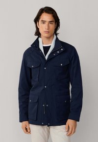 Chaqueta azul marino que presenta dos bolsillos en el pecho, dos bolsillos inferiores y un cuello alto con cierre de cremallera y broche. Hecha de una tela suave y duradera.