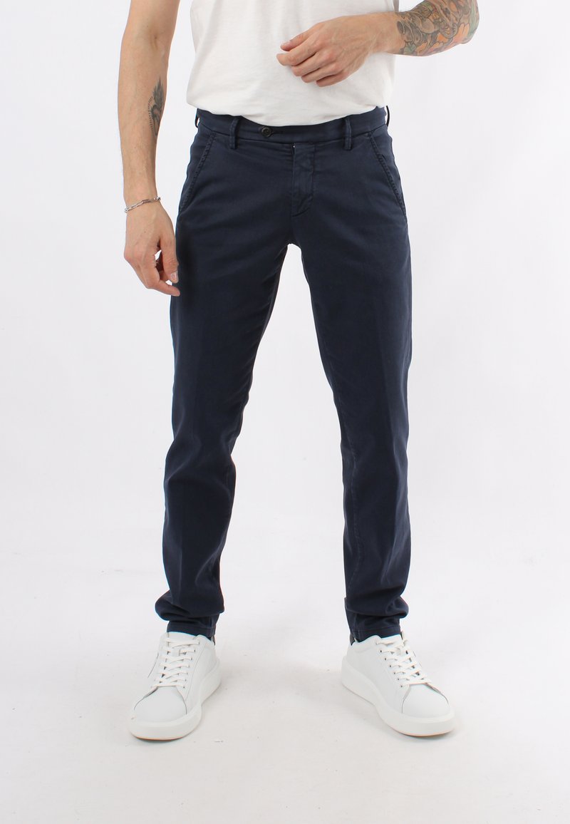 Chino in cotone blu navy con vestibilità slim, tasche frontali e chiusura a bottone, abbinati a sneakers bianche. Texture liscia, dettagli minimi.