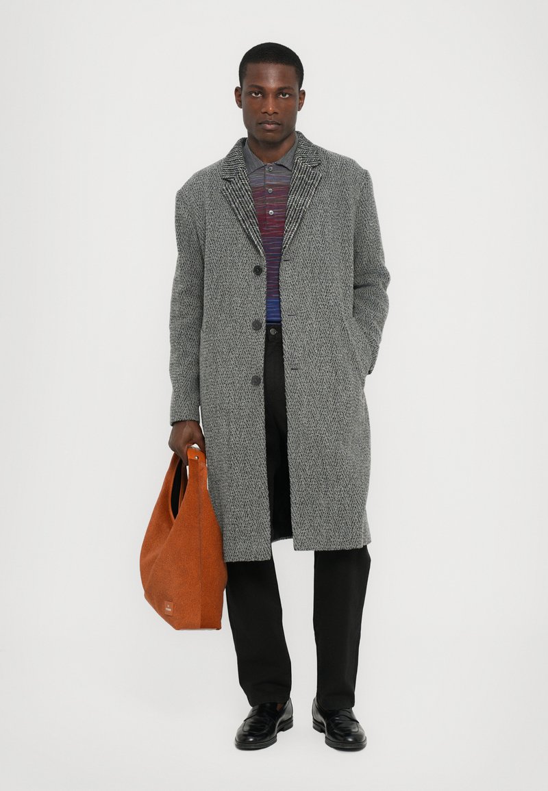 Grijs visgraat jasje met een notchkraag, gecombineerd met een meerkleurig gestreepte polo. Houdt een oranje draagtas vast, met zwarte broek en schoenen.