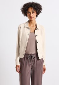 Ljusbeige jacka med krage, knappstängning fram och två sidofickor. Bärs över en taupe-top med löst sittande bruna byxor.