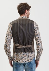 Gilet a righe marroni con un dettaglio a scacchi in vita, indossato sopra una camicia a fantasia floreale con una combinazione di colori beige e nero. Jeans in denim visibili.